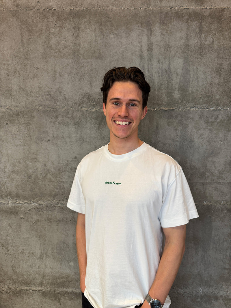 Feder & Kern Mitgründer Cedric in  weißem T-Shirt,  lächelnd vor Betonwand